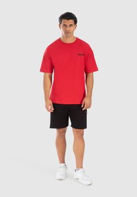 Rød bomulds T-shirt med korte ærmer, med et sort "SMILODON" logo, parret med sorte shorts og hvide sneakers. Enkel baggrund.