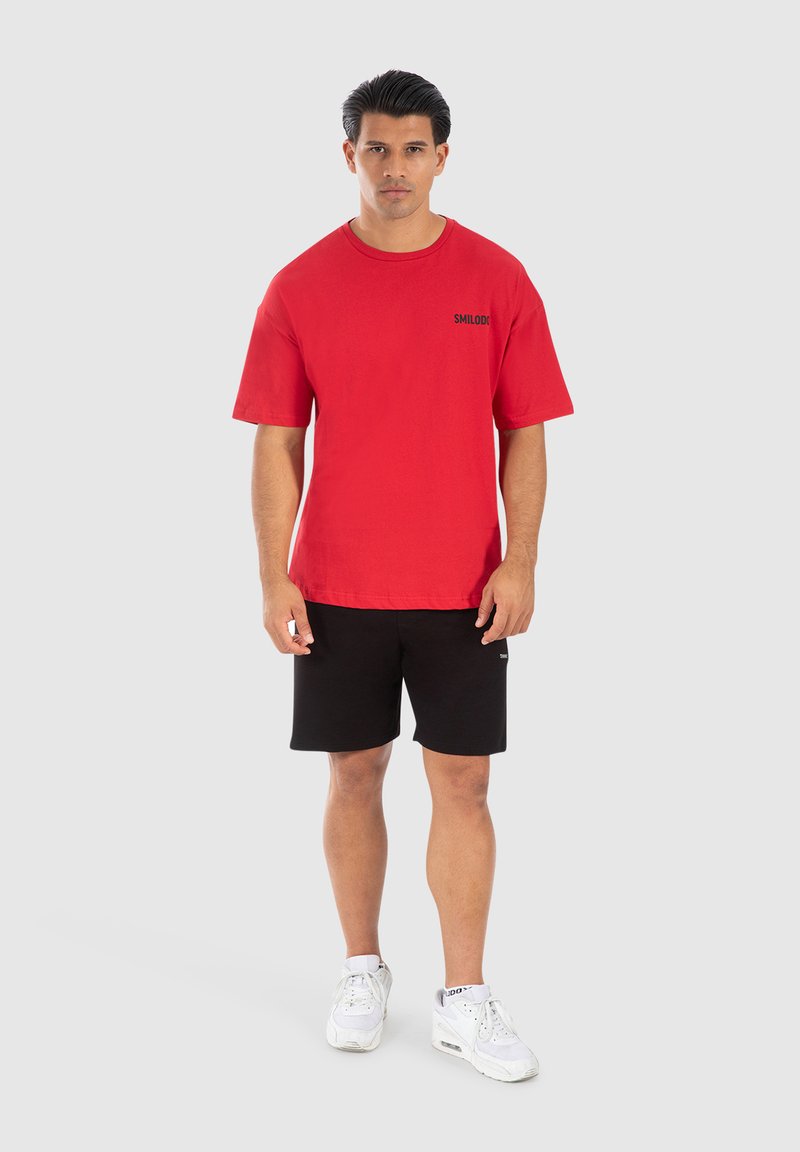 Rød bomulds T-shirt med korte ærmer, med et sort "SMILODON" logo, parret med sorte shorts og hvide sneakers. Enkel baggrund.