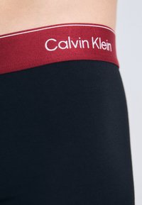 Zwarte boxershorts met een bordeauxrood elastische tailleband waarop "Calvin Klein" in witte tekst staat. Glad textuur en een aansluitende pasvorm.