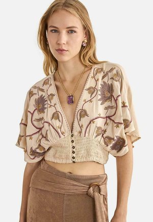 Mujer con blusa corta beige bordada con flores moradas, con botones al frente y mangas amplias, combinada con pantalones marrones de cintura alta con cinturón atado.