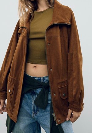 Personne portant une veste en suède marron par-dessus un crop top vert olive et un jean bleu avec un pull noir noué autour de la taille.