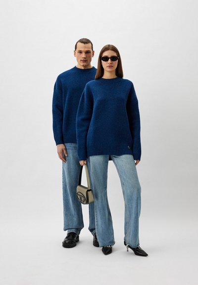 Blaue Strickpullover mit gerippten Bündchen, kombiniert mit hellblauen Jeans. Eine Person hält eine kleine beige Handtasche.
