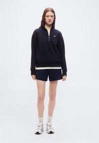 Γυναίκα με μπλε σκούρο φούτερ με φερμουάρ στο ύψος του λαιμού, μπλε σορτς, λευκά αθλητικά παπούτσια και κάλτσες, στέκεται μπροστά σε ένα απλό φωτεινό φόντο.