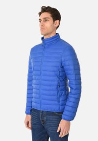 Giacca imbottita blu con colletto alto, zip frontale e tasche laterali. Presenta una texture trapuntata orizzontale e polsini elasticizzati.