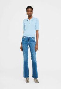 Polo shirt bleu clair avec des manches courtes bouffantes et trois boutons ; associé à un jean bleu évasé avec des accents de bijoux sur les poches.
