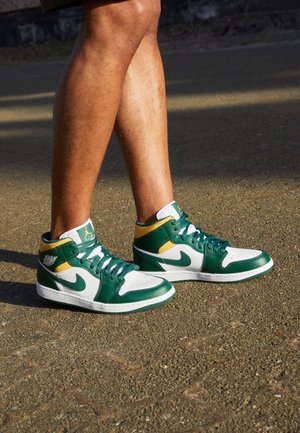 Personne portant des baskets Nike Air Jordan vertes, blanches et jaunes, debout sur un sol en asphalte sous le soleil.