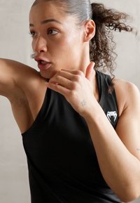 Čierna športová bezrukávová blúzka s logom zobrazujúcim dizajn hada. Kučeravé vlasy čiastočne stiahnuté, ústa mierne otvorené v dynamickej póze.