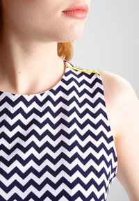 Robe sans manches avec un motif en chevrons bleu marine et blanc, présentant un tissu texturé et un accent jaune vif le long de l'encolure.