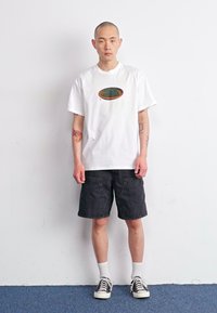 Hvit t-skjorte med et rundt grønt og brunt grafisk motiv, kombinert med svarte denimshorts. Modellen har på seg svart-og-hvitt sneakers med hvite sokker.