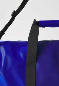Blue waterproof tote bag with a glossy finish, featuring a black adjustable strap, a snap closure, and embossed branding.