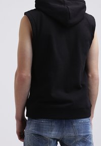 Sudadera sin mangas negra con un tejido texturizado y una gran capucha; presenta un corte relajado y un dobladillo acanalado, combinada con unos pantalones vaqueros de mezclilla azul claro.