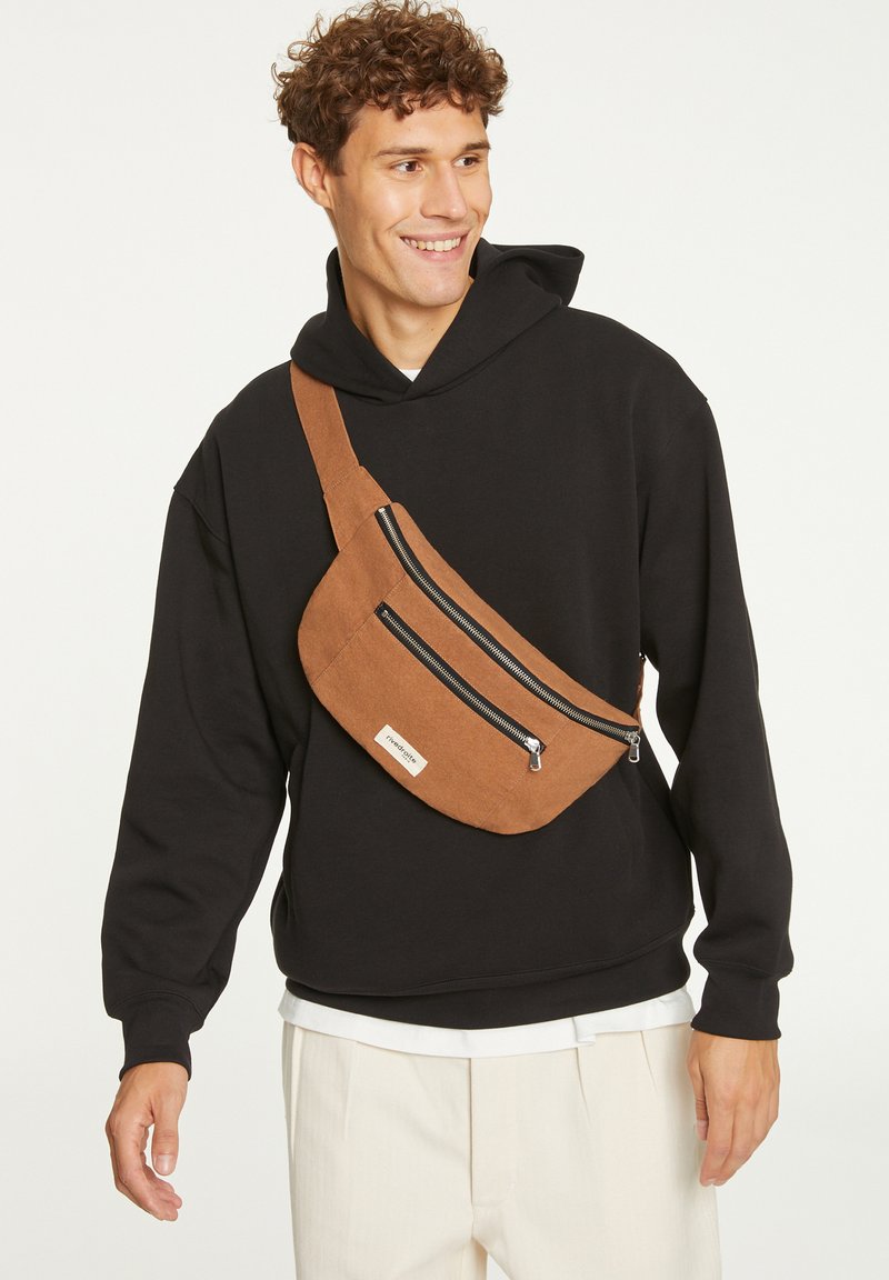 Sac en toile marron en bandoulière avec deux poches à fermeture zippée noires, porté par-dessus un hoodie noir. Le sac présente un design plat et incurvé avec une sangle ajustable.