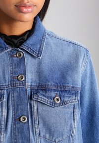 Světle modrá džínová bunda s límečkem, dvěma zapínacími kapsami na hrudi a stříbrným kováním. Má vybledlou texturu a viditelné švy.