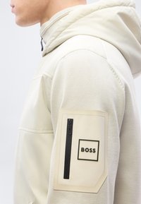 Πλάγια όψη ατόμου που φοράει μπεζ φούτερ με κουκούλα, με τσέπη με φερμουάρ και το λογότυπο "BOSS" στο πάνω μέρος του μανικιού.