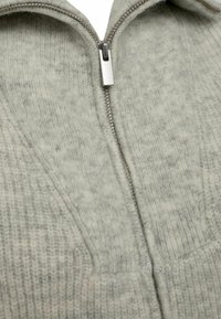 Siv sweater z zadrgo, z teksturirano pleteno strukturo in kapuco. Tkanina je mehka z vidnimi vlakni in subtilnimi barvnimi razlikami.
