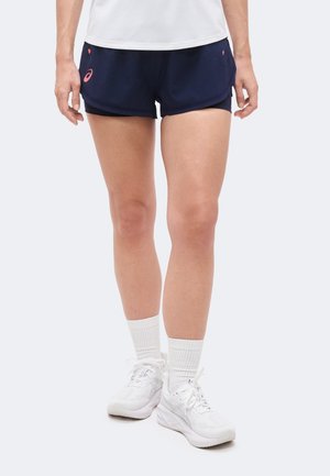 Személy tengerészkék és fekete réteges futónadrágban, fehér bokazokniban és fehér sportcipőben, egyszínű háttér előtt áll.