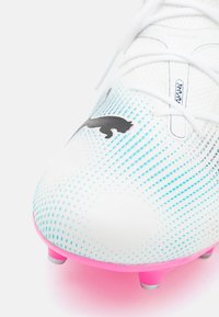 Scarpini da calcio bianchi con motivi a pois blu e un logo nero. Presentano una superficie testurizzata e una suola vibrante rosa con tacchetti visibili.