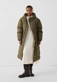 Abrigo acolchado de color verde oliva con capucha, que presenta una silueta larga, bolsillos laterales y cierre con cremallera. Llevado sobre un vestido blanco y botas marrones.