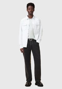 Camicia di denim bianco sopra una maglietta verde chiaro, abbinata a jeans neri a gamba dritta e scarpe nere massicce. L'outfit include una cintura argentata.