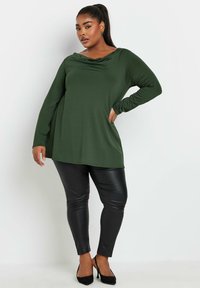 Grön långärmad topp med krage, kombinerad med åtsittande svarta faux läderbyxor och svarta högklackade skor med spetsig tå.