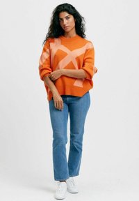 Orange oversized tröja med rosa geometriska mönster, rund halsringning, ribbade ärmslut, tillsammans med ljusblå jeans och vita sneakers.