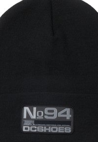 Černá pletená čepice s kulatým vrškem. Obsahuje obdélníkovou šedou nášivku s nápisem "No. 94" a "DC Shoes" bílým písmem.