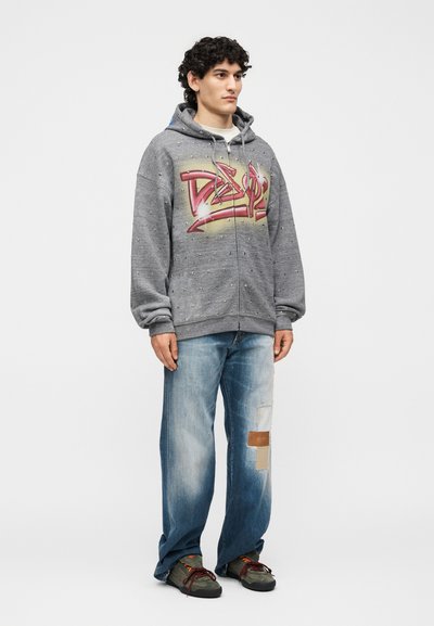 Νέος άνδρας με γκρι φούτερ με φερμουάρ και σχέδιο γκράφιτι, σκισμένα χαλαρά μπλε τζιν με επιδιόρθωση και σκοτεινά αθλητικά παπούτσια, στέκεται μπροστά σε ένα μονόχρωμο φόντο.
