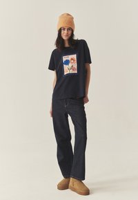 Tumesinine T-särk lillelise trükiga, kombineeritud tumedate teksade ja beeži seemisnahast sussidega. Modell kannab kudumit oranži mütsi.