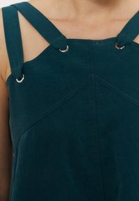 Gros plan d'une personne portant un vêtement sans manches vert sarcelle foncé avec de larges bretelles fixées par des anneaux métalliques aux épaules.