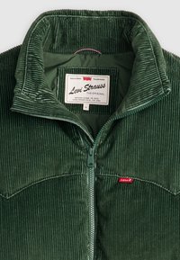 Zielona kurtka sztruksowa z wysokim kołnierzem, zamkiem na pół guzika z przodu i metką Levi's. Charakteryzuje się teksturowaną tkaniną oraz kontrastowym czerwonym logo przy zamku.