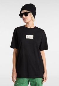 Sort bomulds t-shirt med et hvidt Vans-logo på brystet, parret med grønne cargo-bukser og en sort ribbet beanie. Mørke solbriller bæres.