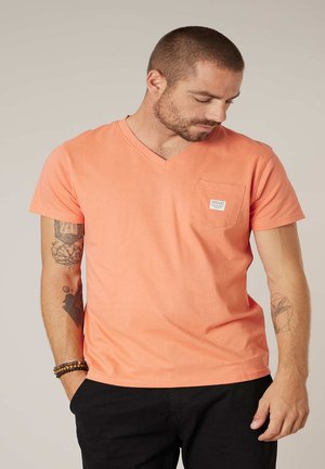 Homme aux cheveux courts et barbe, portant un t-shirt corail à col en V avec une poche sur le devant et un pantalon noir, regardant vers le bas les mains dans les poches.