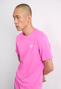 Rausvas medvilninis marškinėliai su apvalia iškirpte ir trumpomis rankovėmis; ant kairės krūtinės yra mažas baltas Nike logotipas; lygi tekstūra.