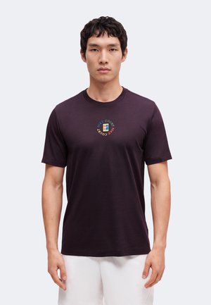 Uomo che indossa una maglietta Nike viola scuro con un logo circolare colorato al centro e pantaloncini bianchi, in piedi davanti a uno sfondo semplice.