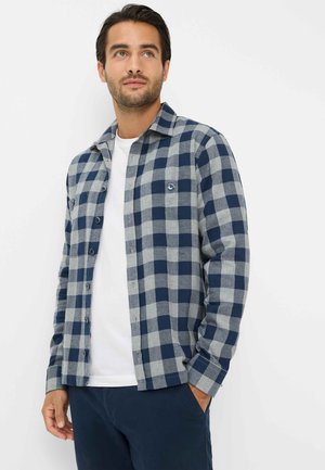 Camicia a scacchi blu navy e grigia chiara con colletto button-down, tasche frontali e logo sul petto, abbinata a una t-shirt bianca a girocollo.