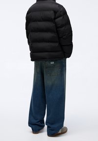 Personne portant une doudoune noire, un jean bleu à jambes larges avec des poches arrière, et des baskets grises, debout devant un fond blanc.