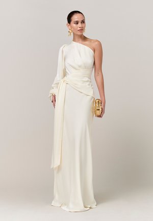 Vrouw draagt een lange, ivoorkleurige jurk met één schouder, houdt een kleine gouden clutch vast en heeft grote gouden oorbellen als accessoires.