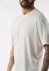 Maglietta bianca a maniche corte realizzata in tessuto morbido, con un logo goffrato sottile sul petto e una vestibilità casual. Texture liscia e design pulito.