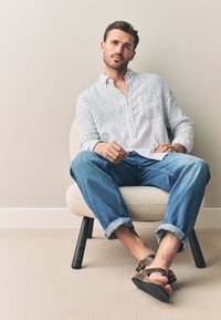 Gestreiftes weißes Hemd mit Knöpfen, blaue hochgekrempelte Jeans und braune Sandalen. Der Stuhl ist rund und mit einem hellen Stoff bezogen, hat schwarze Beine.