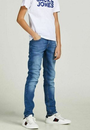 Person iført slidte blå slim jeans, hvid "JACK & JONES" t-shirt og hvide sneakers med mørke striber, stående med den ene hånd i lommen.