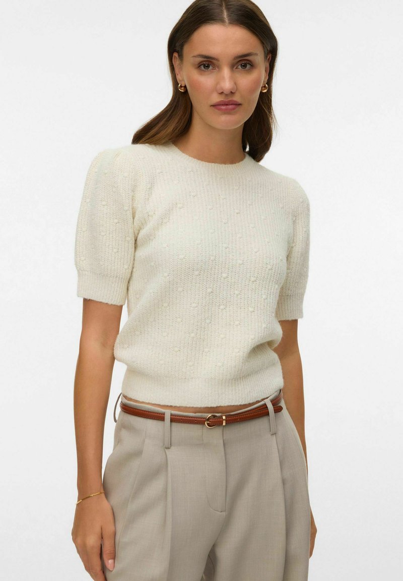 Pull en maille crème avec manches courtes bouffantes, motif texturé. Le mannequin porte un pantalon beige clair avec une ceinture marron. Design simple et décontracté.