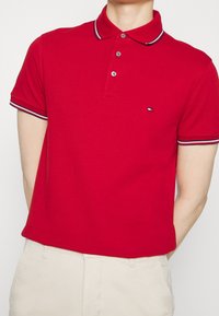 Homme portant un polo rouge à manches courtes avec un col et des manches à rayures blanches, bleues et noires, associé à un pantalon beige.