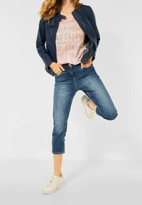 Granatowa kurtka z ekoskóry, jasnoróżowa koszulka z nadrukiem, ciemnoniebieskie krótkie dżinsy i białe trampki. Modelka przyjmuje figlarną pozę.