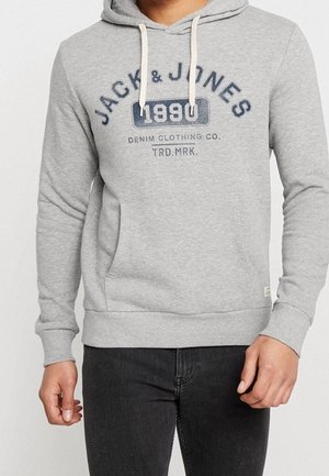 Personne portant un sweat à capuche gris clair avec le texte "JACK & JONES 1990 Denim Clothing Co. TRD. MRK.", associé à un pantalon noir.