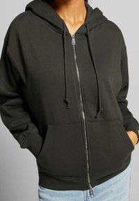 Tummanharmaa vetoketjullinen huppari, joka on valmistettu pehmeästä kankaasta ja jossa on huppu, nyörit ja kenguru-taskut. Ribbaatut hihansuut ja helma.