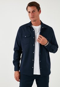 Cămașă cu nasturi de culoare bleumarin, cu un buzunar pe piept, guler clasic și mâneci lungi, realizată dintr-un material neted, purtată peste un tricou alb.