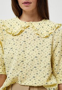 Blusa gialla con texture, colletto arricciato, decorata con un motivo floreale blu e verde. Maniche corte, vestibilità comoda e tessuto morbido.