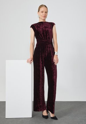 Combinaison en velours bordeaux avec des manches courtes, un détail de taille torsadé et des jambes larges, associée à des talons noirs à bout pointu et à un bracelet en argent.