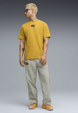 Junger Mann mit kurzen blonden Haaren und Sonnenbrille trägt ein senfgelbes T-Shirt, hellgraue Hose mit Aufnähern und gelbe Turnschuhe, steht vor einem grauen Hintergrund.