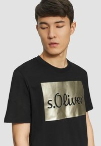 Schwarzes Baumwoll-T-Shirt mit einem großen, metallisch goldenen rechteckigen Akzent, auf dem der Text "s.Oliver" in fetter schwarzer Schrift steht. Mit kurzen Ärmeln und rundem Halsausschnitt.